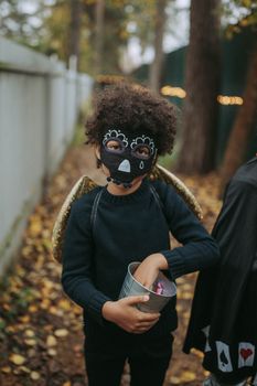 一个孩子穿着有趣的万圣节服装，戴着面具和翅膀，手里拿着糖果桶