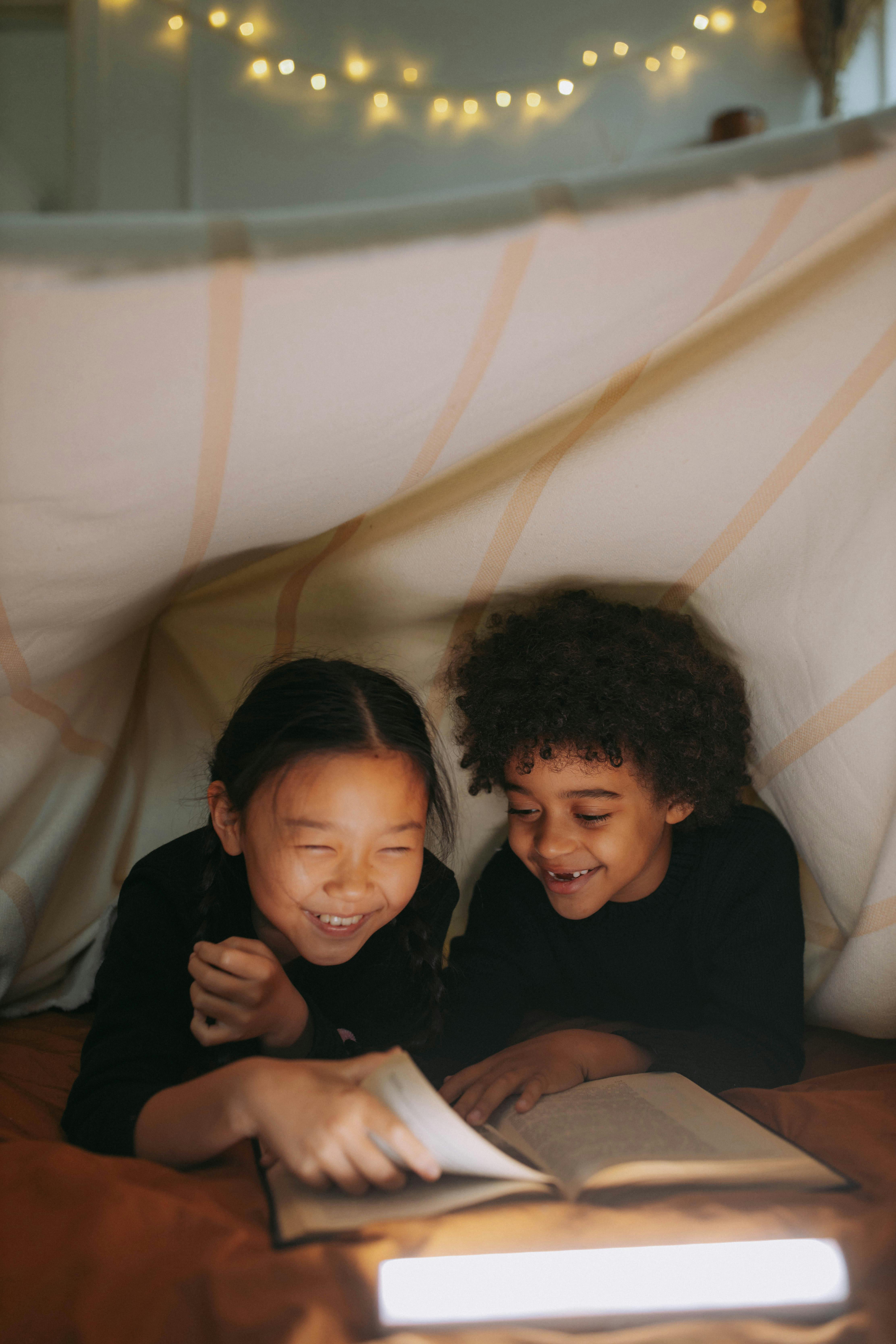 a young girl and a boy reading a book while under the blanket