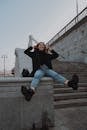 From below of young cheerful female in stylish wear having fun on stone fence while touching hair near city staircase