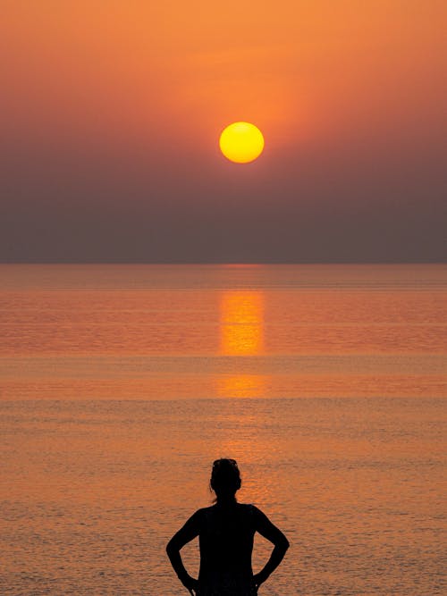 Back view of female observing bright tranquil sunset reflecting in calm rippling water of sea
