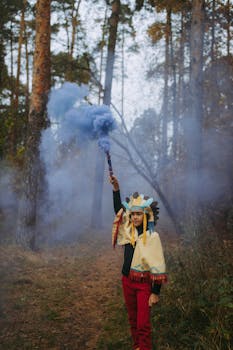 在宁静的森林里，一名穿着文化服装的年轻人站在高大的树木间，手持蓝色烟雾弹