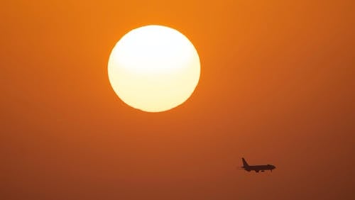 Silhouette of airplane flying in cloudless orange sky under bright sun in sunset time