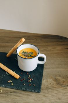 Cozy autumn pumpkin soup in white enamel mug with breadsticks on wooden table.