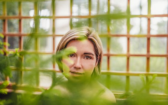 Through green plant branches pretty young female with bare shoulders and short haircut standing in garden and looking at camera dreamily