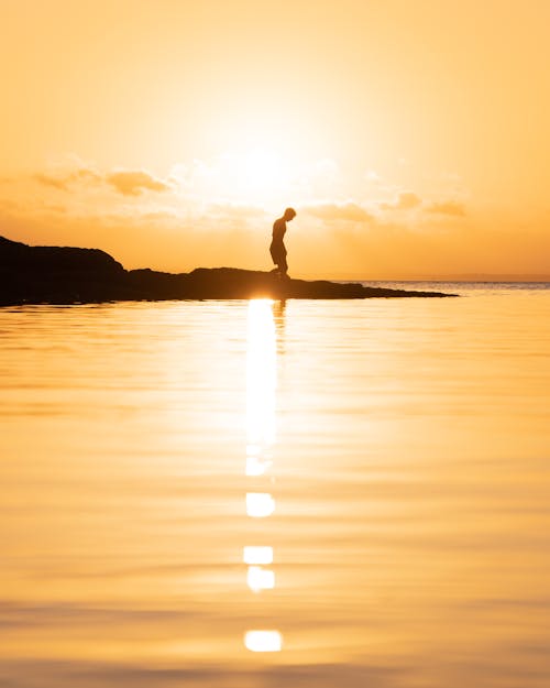 Silhouette of a Man on a Rock Amid the Ocean Water Golden From the Light of Setting Sun