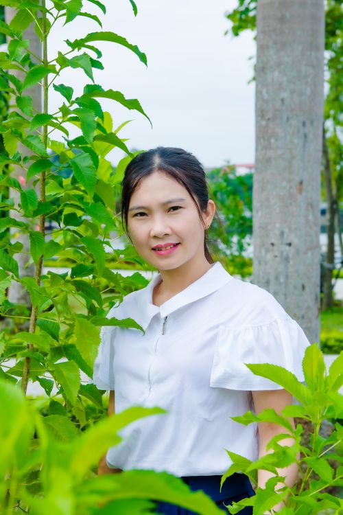 Free Asian woman in a white blouse standing in a lush garden, surrounded by trees. Stock Photo