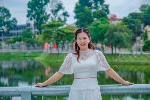 Free Elegant portrait of a woman in a white dress standing by a river with a lush green backdrop. Stock Photo