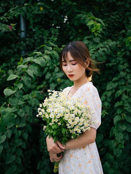 一位迷人的年轻女子闭眼站在灌木旁，手持盛开的花朵，呈现侧面轮廓