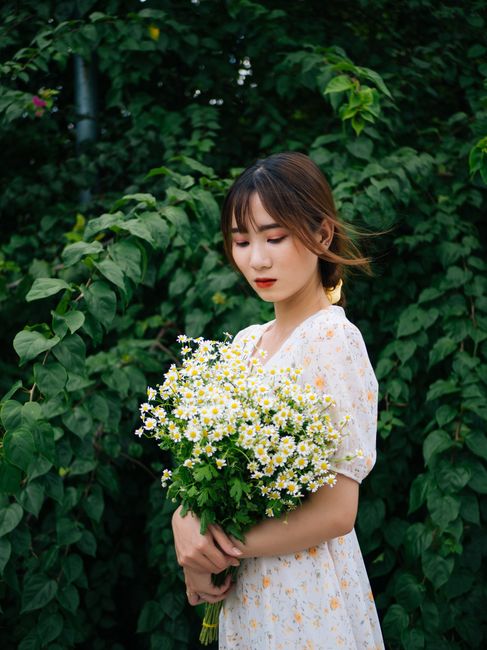 Side view of attractive young female standing with closed eyes and holding bouquet of blooming flowers near shrubs