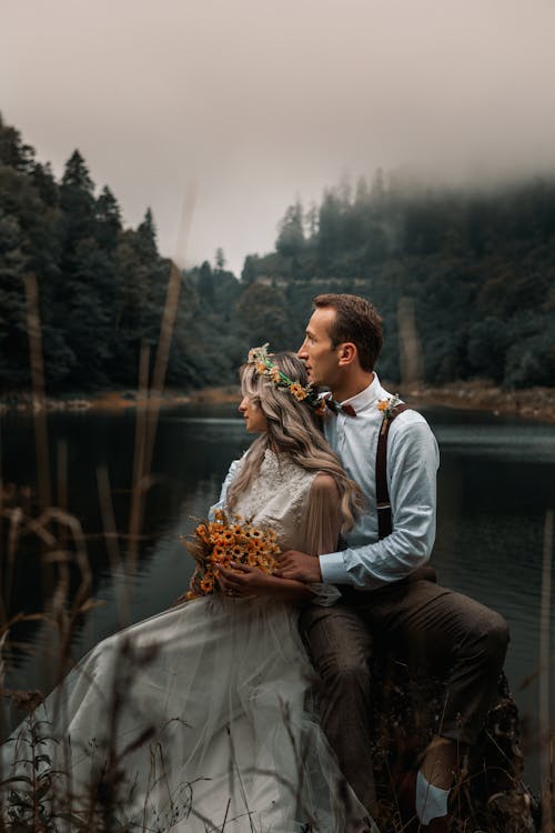 Young groom gently embracing bride with flower wreath and bouquet while sitting on river shore against woods and looking away