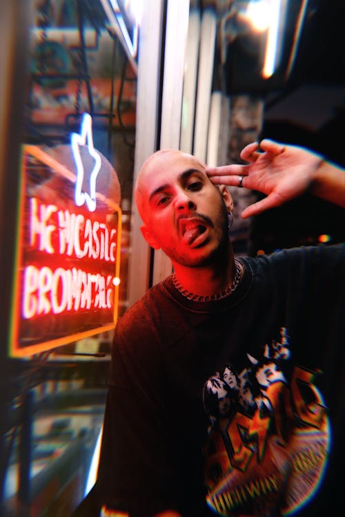A Man Beside a Neon Signage with a Finger Gun on Head