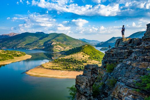 A solitary hiker stands on a rocky cliff overlooking a peaceful mountain lake under a blue sky.