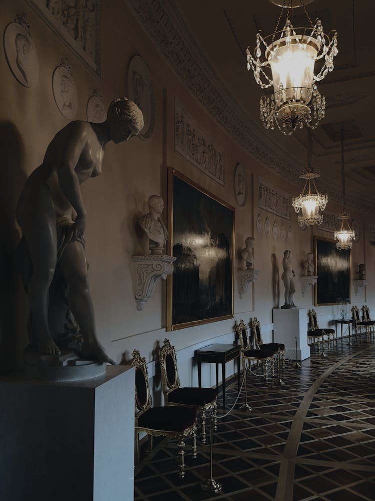 A Dark Museum With Lighted Chandeliers Over Statues And Chairs