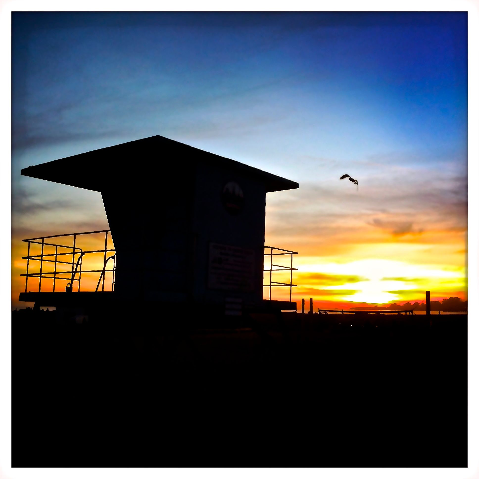 Dramatic silhouette of a lifeguard tower against a vibrant sunset sky with a bird in flight.