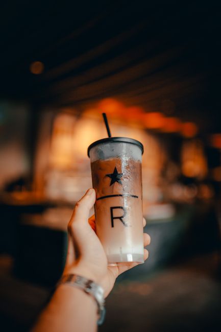 Reusable plastic glass of cold iced coffee with straw in hand of crop unrecognizable person on blurred cafe background