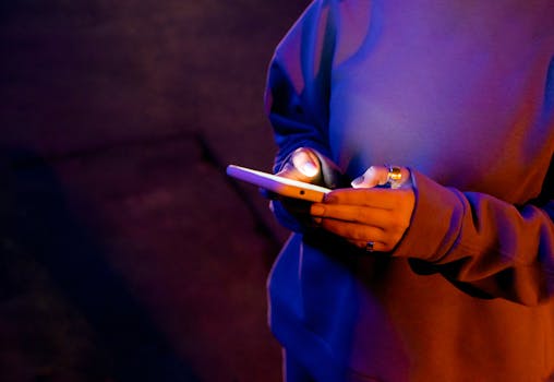 A person in a sweatshirt using a smartphone at night illuminated by screen light.