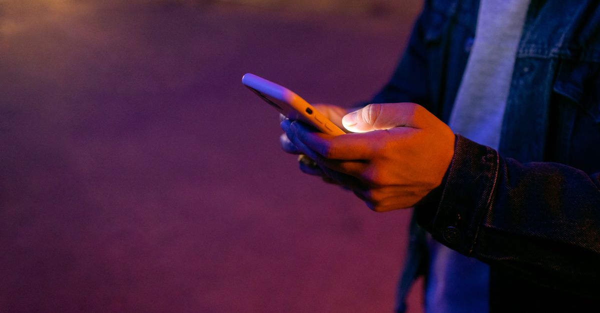 A hand holding a smartphone in a dark room with a neon sign reading "HOTEL CALIFORNIA"