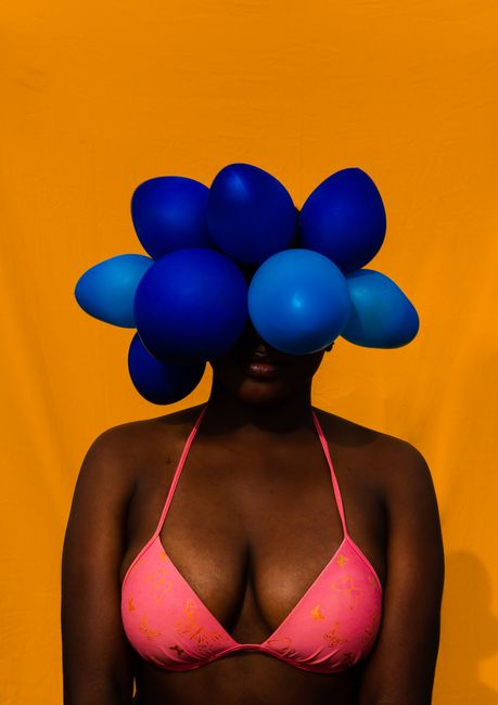 Anonymous carefree African American female wearing pink bikini standing on bright orange background with blue balloons on head covering eyes