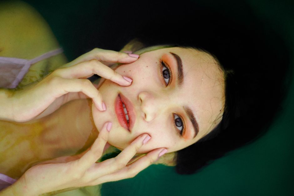 From above of dreamy young ethnic female with dark hair and makeup touching face and looking at camera with mouth opened while resting in reservoir