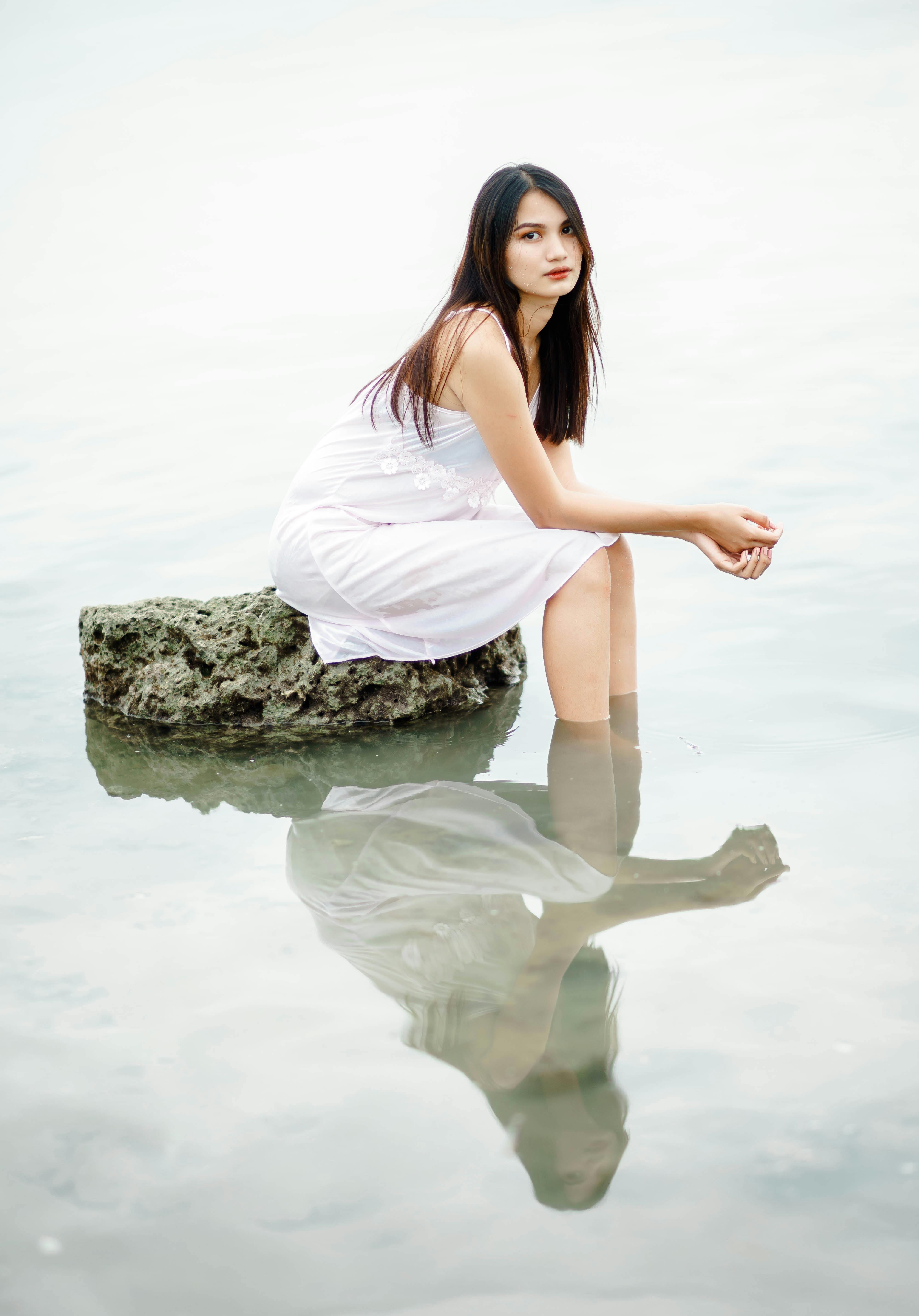 Beautiful young slender female in white dress looking at camera and reflecting in river while resting on large stone