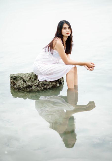 Beautiful young slender female in white dress looking at camera and reflecting in river while resting on large stone