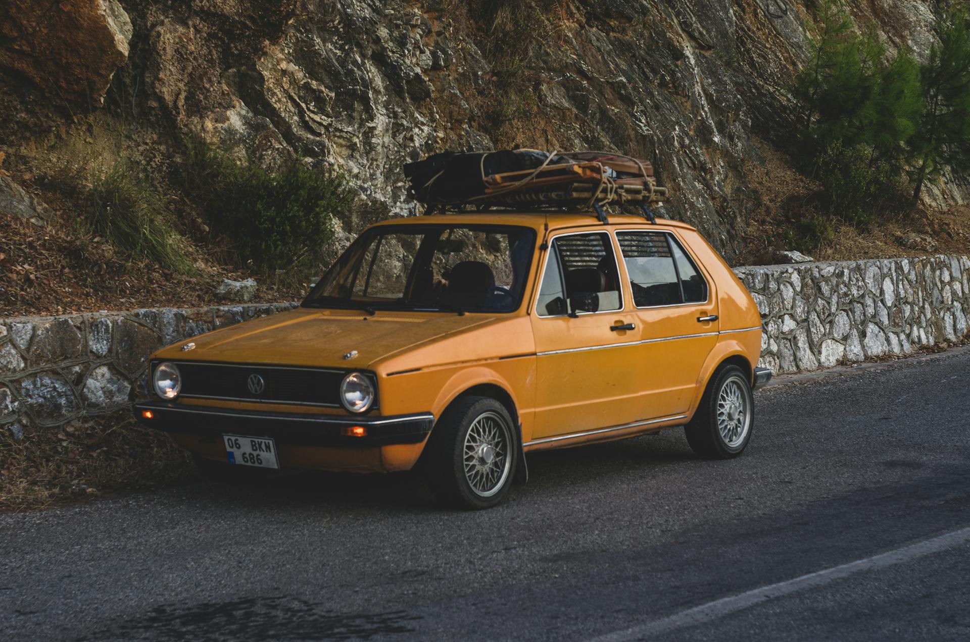 A vintage yellow car with luggage on the roof parked by a rocky mountain road.