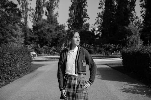 Free Grayscale Photo of Woman in Black Cardigan and Plaid Skirt Standing on Road Stock Photo