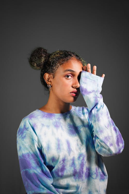Free Attractive young African American female in casual wear covering eye with hand while standing against gray wall and looking at camera Stock Photo