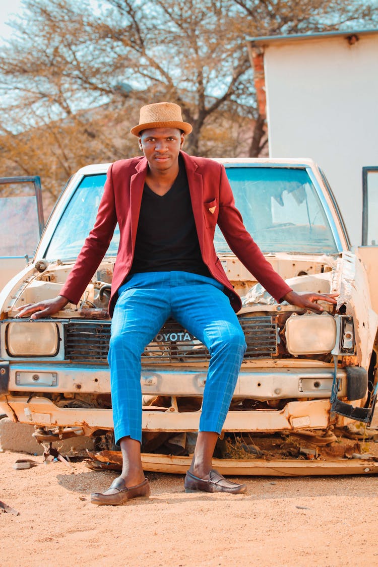 Stylish Black Man Sitting On Hood Of Old Destructed Car In Village