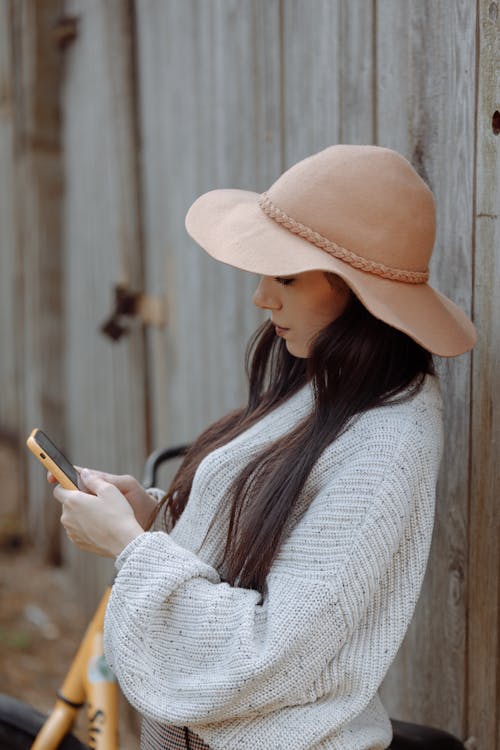 Free Женщина в белом вязаном свитере и бежевой шляпе держит смартфон Stock Photo
