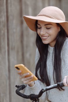 Smiling woman with a smartphone on a bike outside, enjoying a casual autumn day.