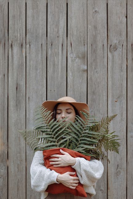 A woman in a hat hugs a fern plant, standing against a rustic wooden wall, conveying nature and tranquility.