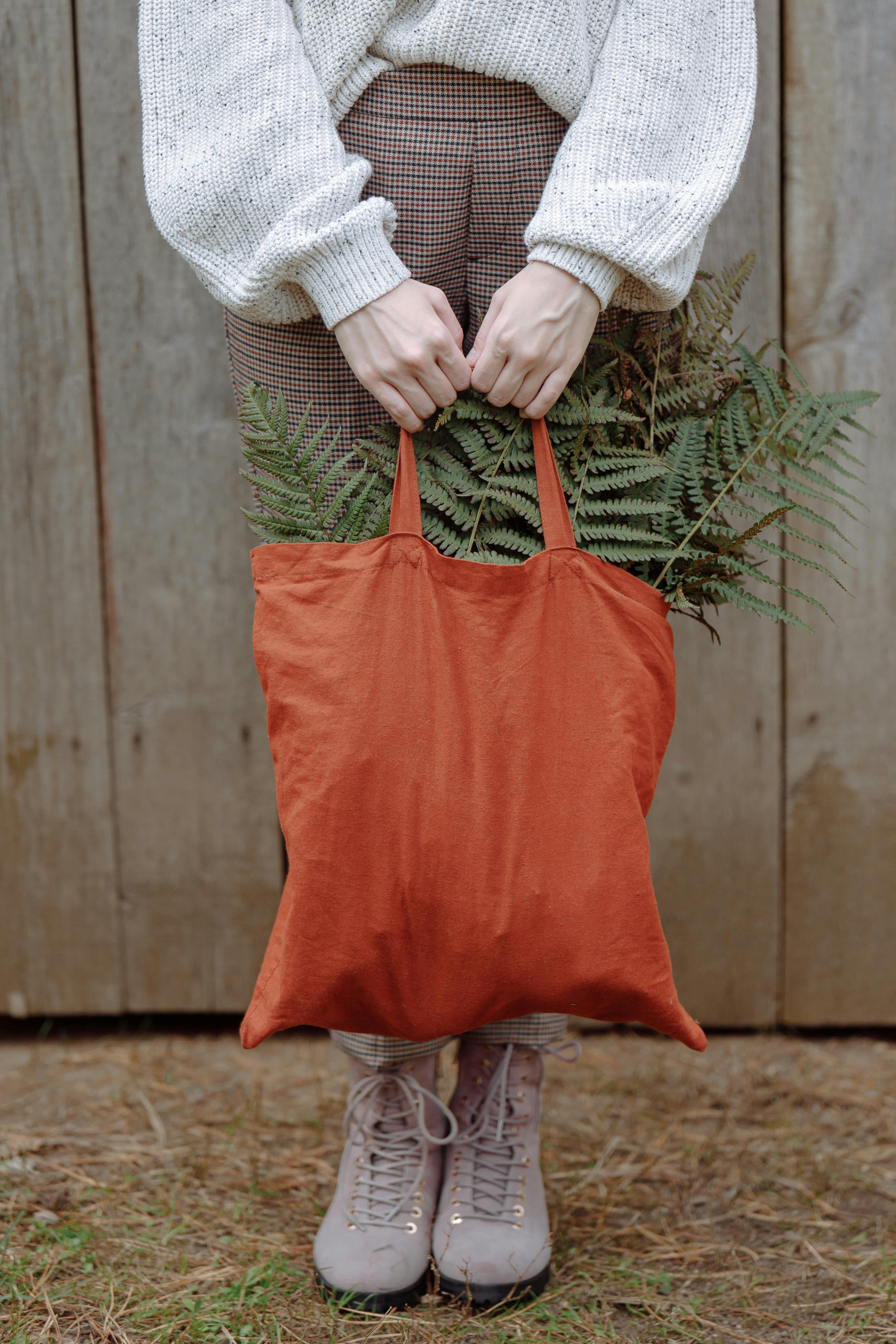Person holding orange bag with ferns, showcasing cozy fall fashion against rustic backdrop.