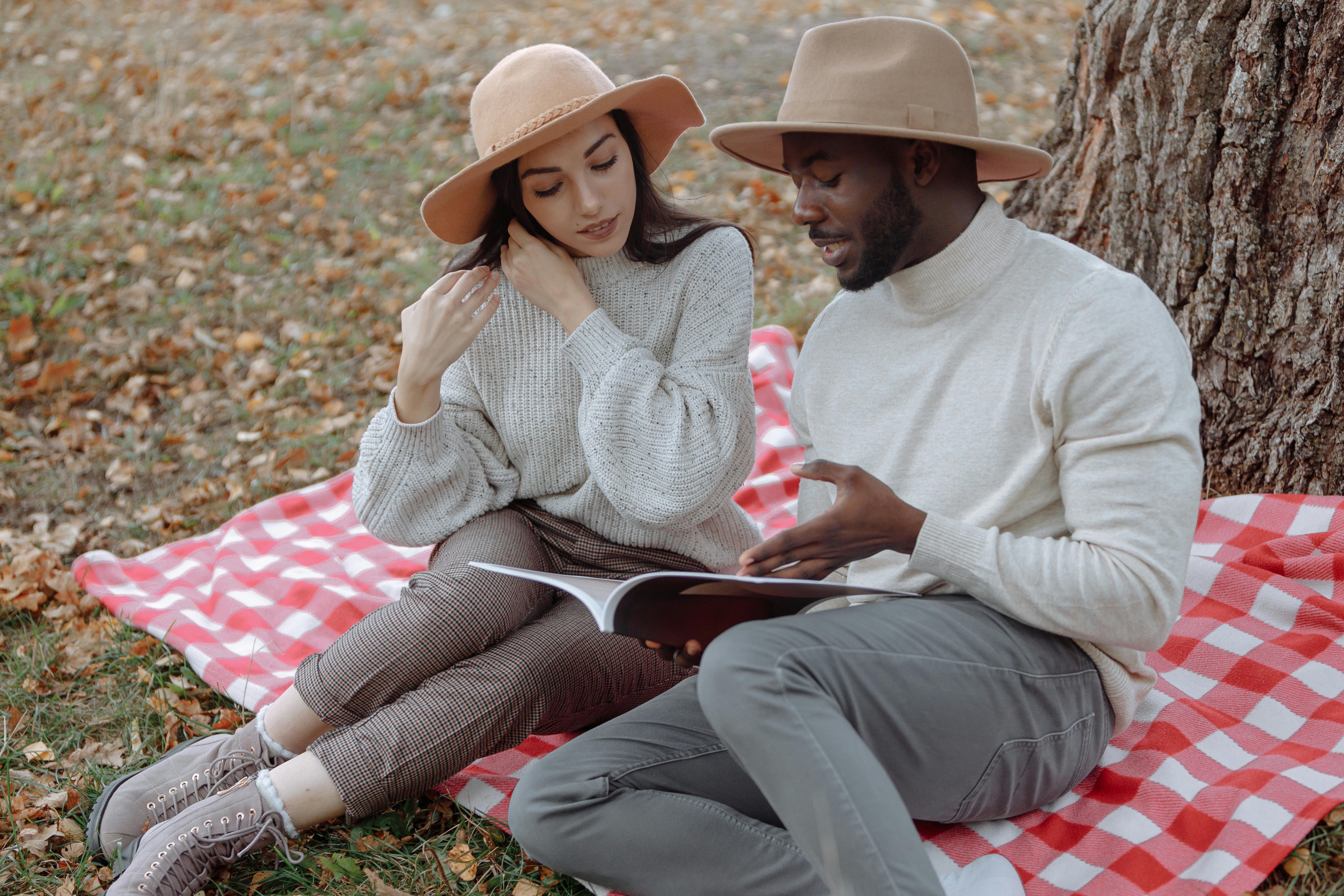 A couple enjoying a picnic in autumn, reading together on a blanket under a tree.