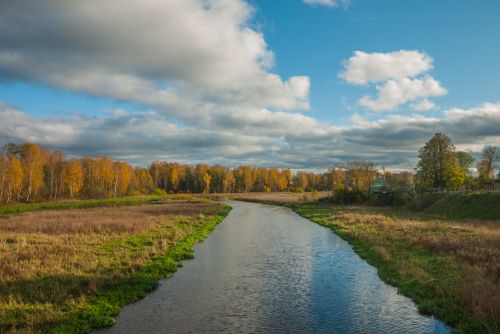 Free Tranquil autumn scene featuring a river, trees with fall foliage, and clouds in the sky. Stock Photo