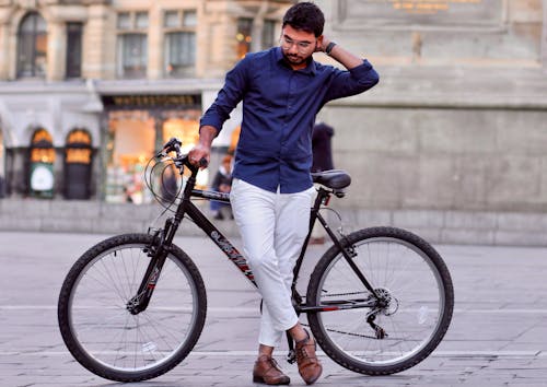 Free Man in Blue Dress Shirt and White Pants Standing Beside A Black Bicycle Stock Photo
