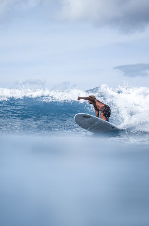 Side view unrecognizable ethnic male surfer balancing on surfboard against foamy sea waves under cloudless sky