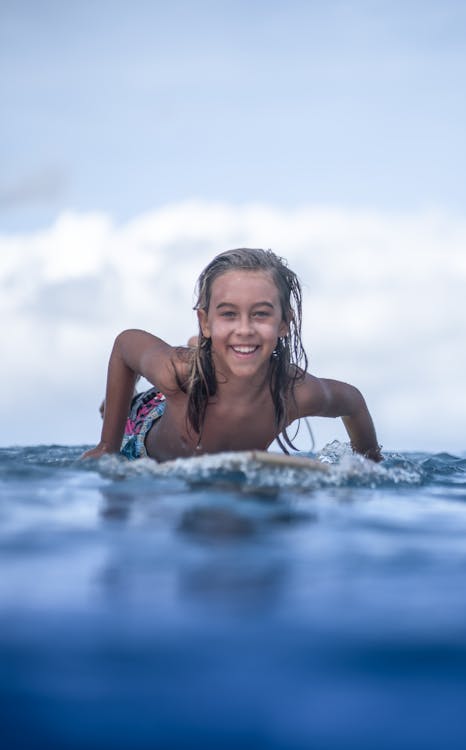 Cheerful teenage boy with long blond hair lying on surfboard on azure seawater and looking at camera with toothy smile