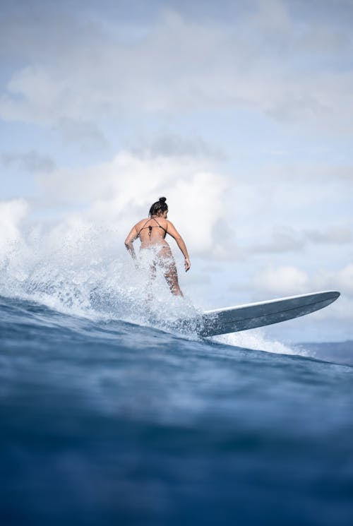 Back view anonymous fit female surfer in black bikini splashing waves on surfboard beneath clear blue sky