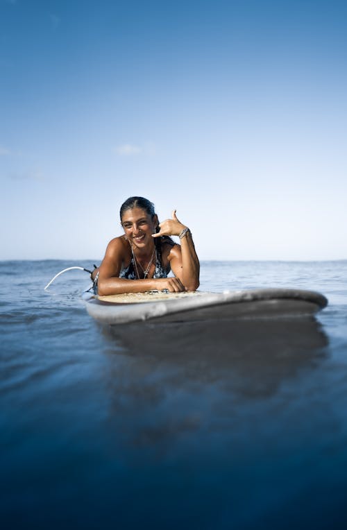 Joyful ethnic female lying on surfboard on blue seawater and showing shaka gesture while looking at camera with toothy smile on clear sunny day