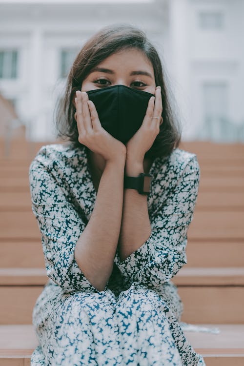 Young calm brunette in casual dress covering face with protective mask while sitting on wooden stairs on street and holding face in hands while looking at camera