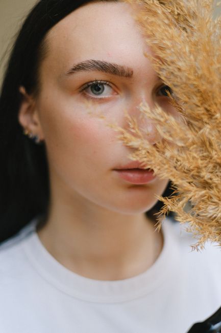 Charming female with dark hair in casual clothes covering face with twig of dried plant and looking at camera in daylight