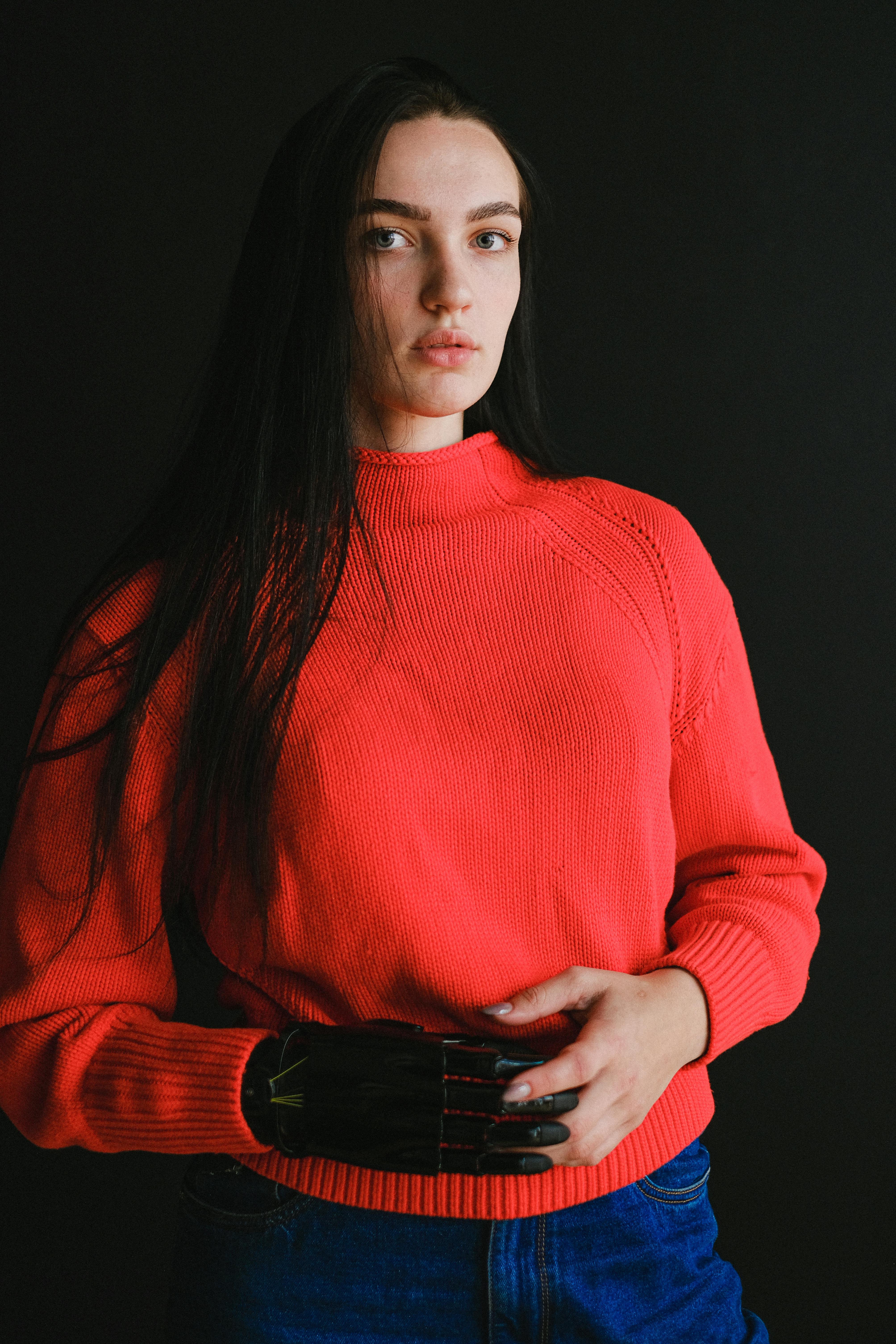 一名女子穿着红色毛衣，在黑暗背景下用仿生假手摆姿势