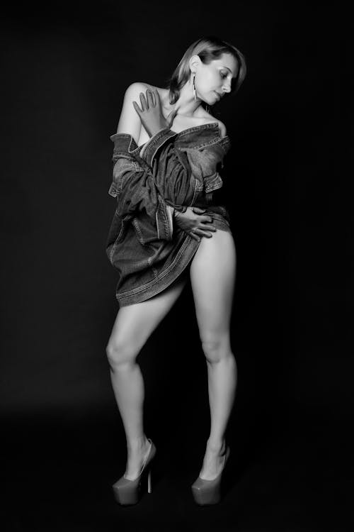 Full body of black and white sensual young lady in stylish denim jacket and high heeled shoes embracing self and looking away in dark studio
