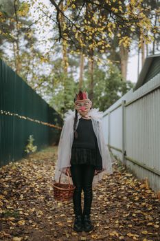 一个穿着万圣节服装的孩子，戴着口罩、提着篮子，站在秋叶中