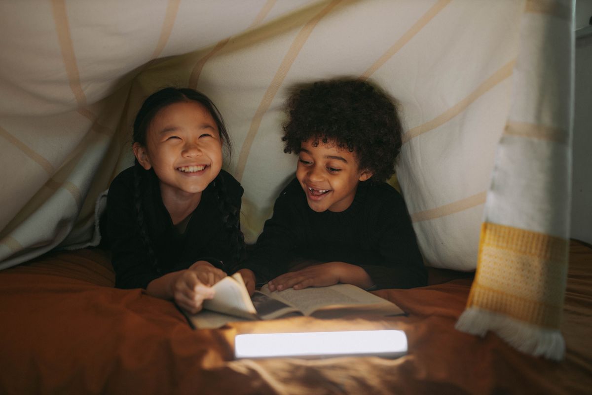 Two children reading a book together in a cozy blanket fort.