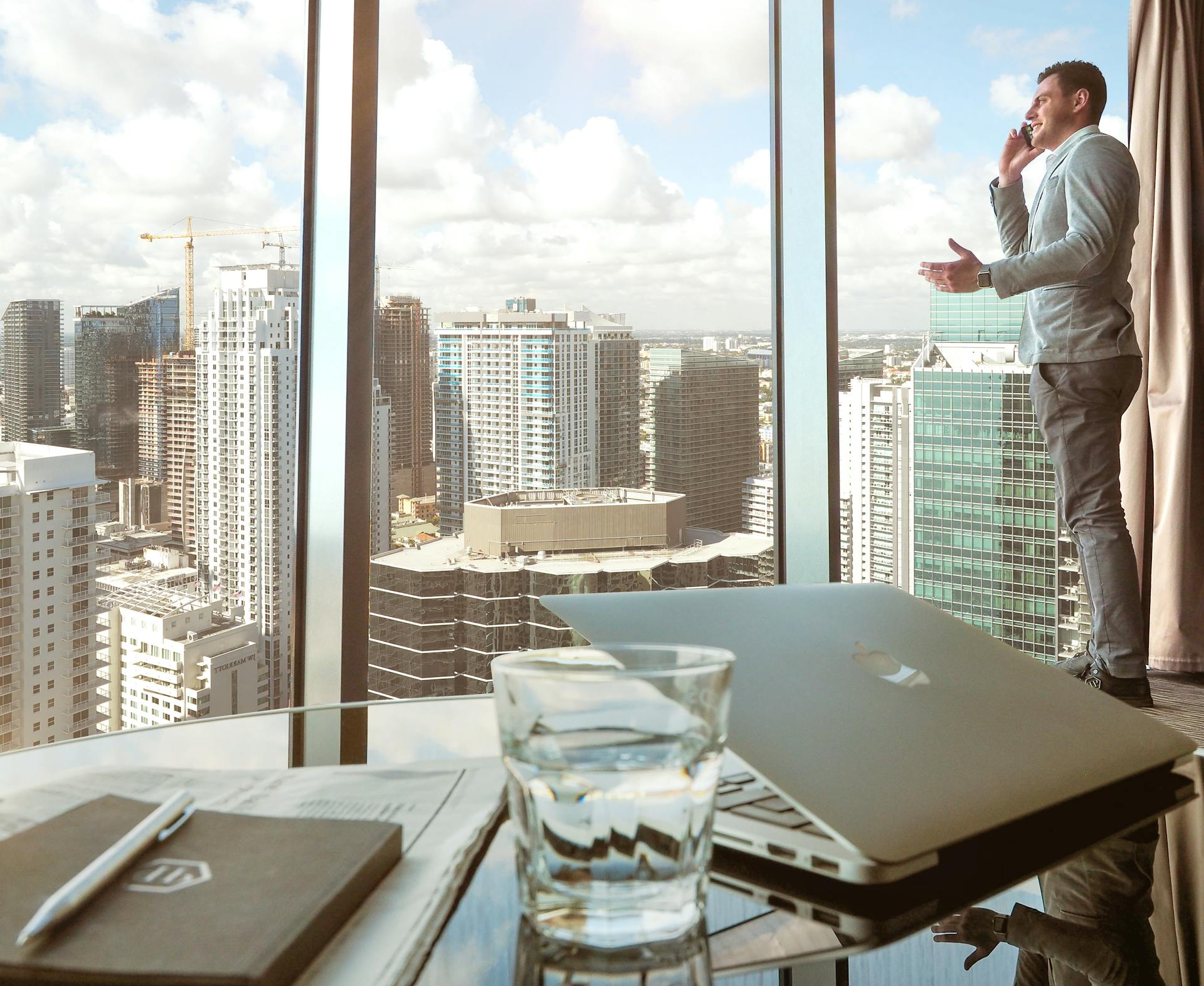 Businessman talking on smartphone by window with city skyline view, laptop and glass in foreground.