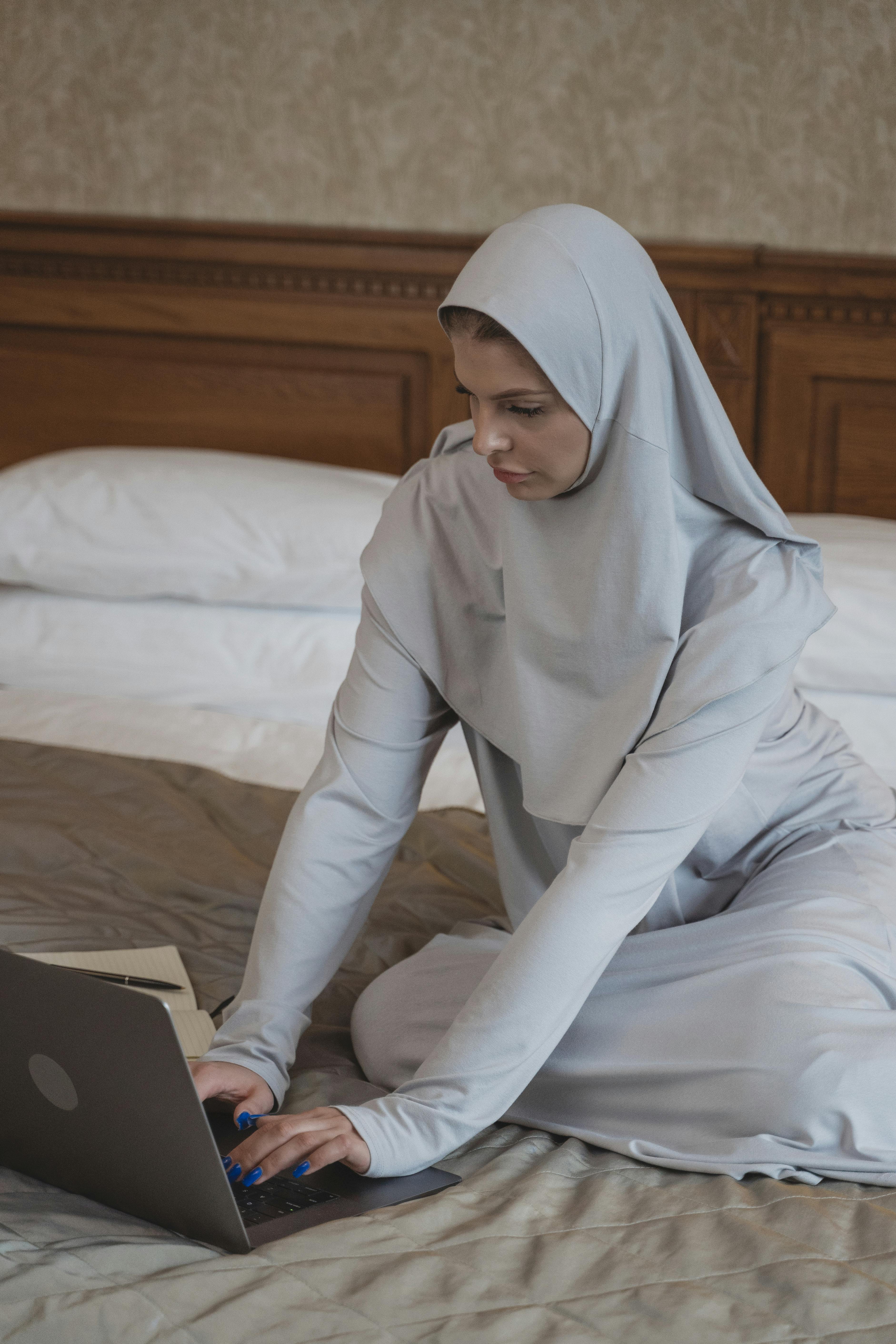 A woman in a hijab sits on a bed, typing on a laptop in a cozy bedroom setting.