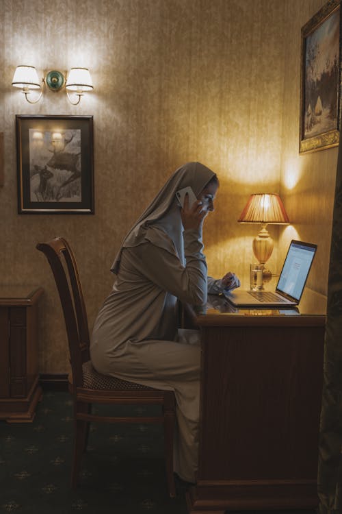 Woman in White Hijab Sitting on Brown Wooden Chair Using Cellphone