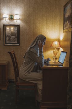 Muslim woman in hijab speaks on phone while working on laptop at home desk, evening light.